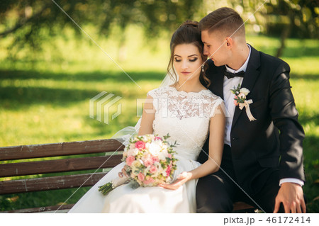 A young couple in wedding clothes is sitting on a bench in a park. Sunny weather A young couple in wedding clothes is sitting on a bench in a park. Sunny weather 46172414