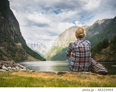 A woman sits on the shore of a picturesque fjord in Norway, admiring the beauty of Scandinavian 46329464