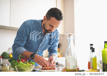 Bearded young man cooking meat while standing near kitchen table Bearded young man cooking meat while standing near kitchen table 46480937