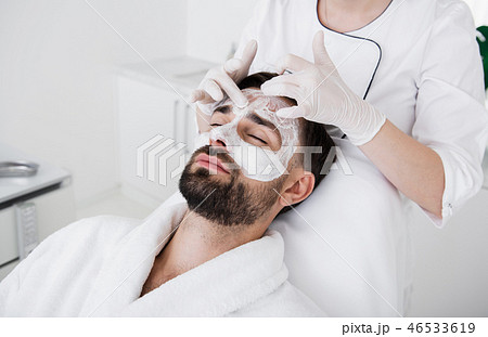 Careful cosmetologist touching white clay mask on the face of man Careful cosmetologist touching white clay mask on the face of man 46533619