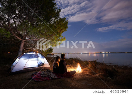 Young couple man and woman having rest at tourist tent and burning campfire on sea shore near forest Young couple man and woman having rest at tourist tent and burning campfire on sea shore near forest 46539944