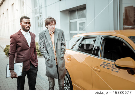 Young student wearing grey coat standing near his student driver car Young student wearing grey coat standing near his student driver car 46830677
