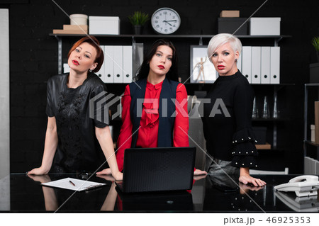 Three business women stand near Desk in office Three business women stand near Desk in office 46925353