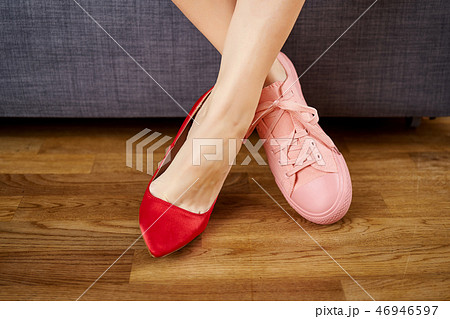 Woman's legs in two different shoes with red high heels and coral sneakers on gray couch background. 46946597