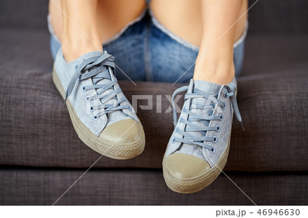View of sexy female legs in jeans shorts and blue sneakers on a gray couch. 46946630