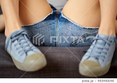 View of sexy female legs in jeans shorts and blue sneakers on a gray couch. View of sexy female legs in jeans shorts and blue sneakers on a gray couch. 46946646