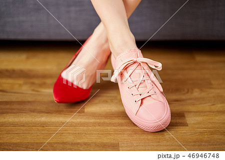 Woman's legs in two different shoes with red high heels and coral sneakers on gray couch background. Woman's legs in two different shoes with red high heels and coral sneakers on gray couch background. 46946748