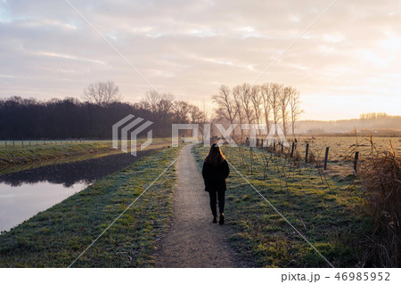 Young woman walks on a cold day by the river in the sunset, beautiful landscape background colorful Young woman walks on a cold day by the river in the sunset, beautiful landscape background colorful 46985952