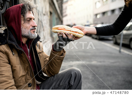 Unrecognizable woman giving food to homeless beggar man sitting in city. 47148780