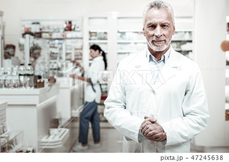 Bearded pharmacist wearing white coat looking pleasant meeting his clients Bearded pharmacist wearing white coat looking pleasant meeting his clients 47245638