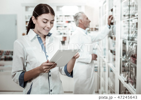 Beautiful dark-haired pharmacist holding tablet while looking at medicine range 47245643