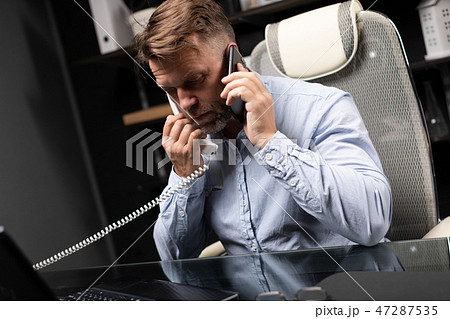young man sitting at computer Desk and holding landline phone and mobile phone young man sitting at computer Desk and holding landline phone and mobile phone 47287535