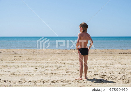 Six-year-old boy in swimming trunks stands on the beach and looks at sea. Six-year-old boy in swimming trunks stands on the beach and looks at sea. 47390449