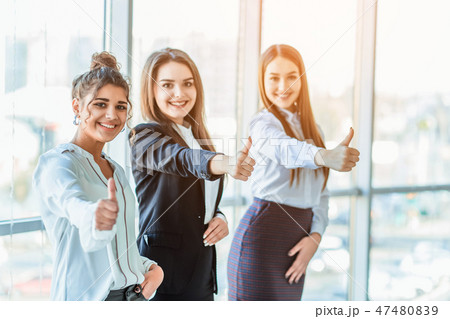 Three young business girls in the office. Dressed in classic clothing style, showing thumbs up 47480839