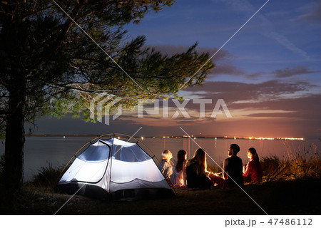 Night summer camping on shore. Group of young tourists around campfire near tent under evening sky Night summer camping on shore. Group of young tourists around campfire near tent under evening sky 47486112