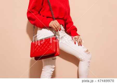 Fashion young woman in white jeans and sweater with red bag clutch in hands near street wall Fashion young woman in white jeans and sweater with red bag clutch in hands near street wall 47489970