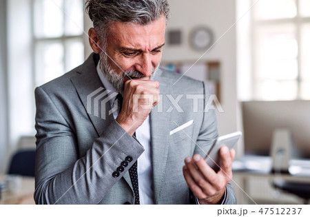 A frustrated businessman with smartphone standing in an office, reading bad news. A frustrated businessman with smartphone standing in an office, reading bad news. 47512237