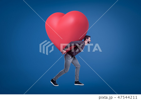 A side view of a young handsome man in a casual outfit carrying a huge red heart on his back. 47544211