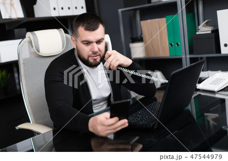 Busy young man talking on phone and looking at laptop in office Busy young man talking on phone and looking at laptop in office 47544979
