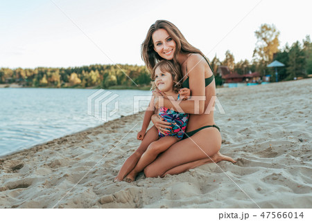 Beautiful and happy young woman mom hugging on shore of lake with sand, little child daughter, happy 47566014