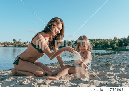 Beautiful young woman mom playing on shore of a lake with sand, next to a small child is a daughter Beautiful young woman mom playing on shore of a lake with sand, next to a small child is a daughter 47566034