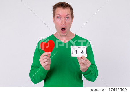 Shocked young man holding calendar block and heart ready for Valentine's day 47642500