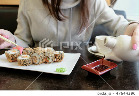 Woman's hand pours soy sauce in small rectangular plate, rolls and cup of tea on background. 47644290