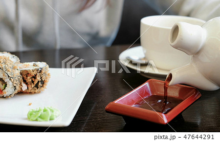 Woman's hand pours soy sauce in small rectangular plate, rolls and cup of tea on background. 47644291