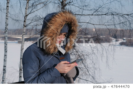 Woman in a blue down jacket with a fur hood writes messaging in her cellphone in a winter Park. 47644470