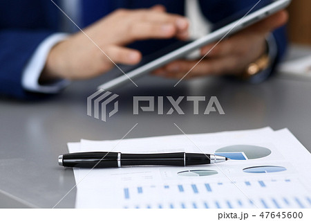 Black pen lying over business analytics papers. Businessman hands using tablet at the background Black pen lying over business analytics papers. Businessman hands using tablet at the background 47645600