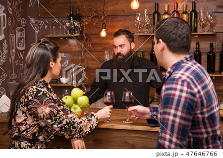 Young barman standing at bar counter talking with girl and her boyfriend. Young barman standing at bar counter talking with girl and her boyfriend. 47646766