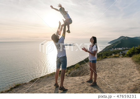 Dad throws his son in the air, and mom looks at them on the mountain with seascape. Family trip. 47659231