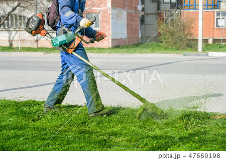 Man wearing overalls and rubber boots mowing green grass with gasoline lawn trimmer. Man wearing overalls and rubber boots mowing green grass with gasoline lawn trimmer. 47660198