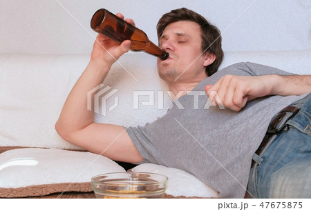 Young man is browsing internet in his mobile phone, eating chips and drinking beer laying on the Young man is browsing internet in his mobile phone, eating chips and drinking beer laying on the 47675875