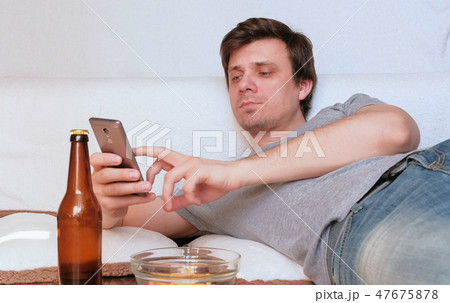 Young man is browsing internet in his mobile phone, eating chips and drinking beer laying on the Young man is browsing internet in his mobile phone, eating chips and drinking beer laying on the 47675878