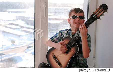 Playing a musical instrument. Boy in sunglasses plays the guitar sitting on the windowsill. Playing a musical instrument. Boy in sunglasses plays the guitar sitting on the windowsill. 47680913