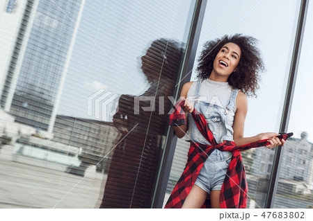 Young woman in the city street near window holding jacket sleeves looking aside excited Young woman in the city street near window holding jacket sleeves looking aside excited 47683802