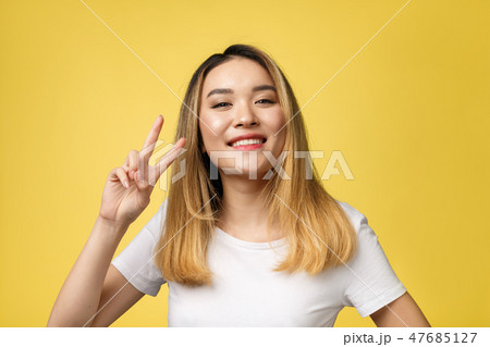 Pleased asian woman in t-shirt showing peace gestures and looking at the camera over yellow Pleased asian woman in t-shirt showing peace gestures and looking at the camera over yellow 47685127