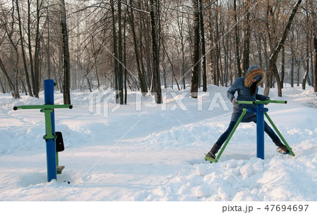 Woman in blue down jacket with hood is doing legs exercises on simulator in a winter Park Front view 47694697