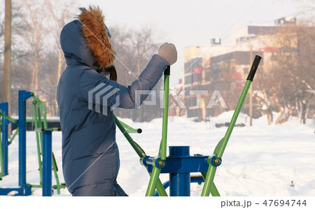 Woman in a blue down jacket with a hood is engaged in ski simulator in a winter Park in city. Woman in a blue down jacket with a hood is engaged in ski simulator in a winter Park in city. 47694744
