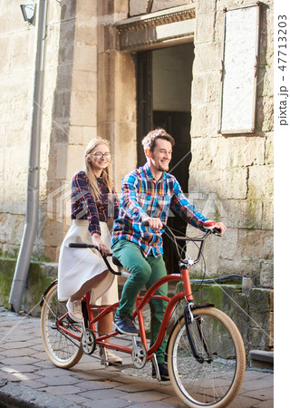 Young tourist couple, handsome man and pretty blond woman riding tandem bicycle along city street. 47713203