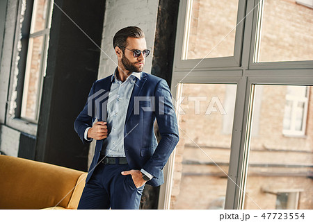 Success concept. Stylish young smiling bearded brunette in sunglasses is standing at office and 47723554