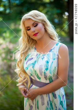 Young beautiful blonde woman in white dress with blue and green flowers posing in forest 47748099
