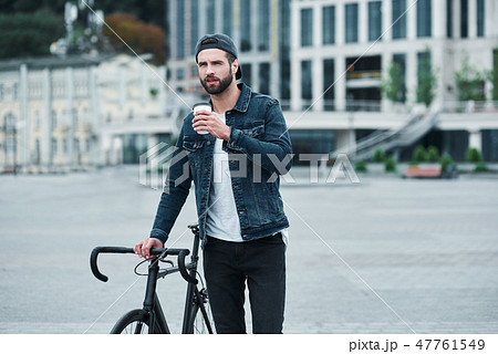 Outdoors leisure. Young stylish man walking on city street with bicycle holding cup drinking hot Outdoors leisure. Young stylish man walking on city street with bicycle holding cup drinking hot 47761549