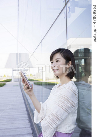 ガラスの壁に寄り掛かってスマホを見る女子大生の写真素材