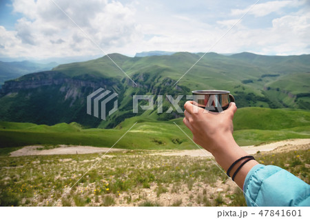 First-person view of a girl's hand holding a plastic cup of tea against a plateau of green hills and 47841601
