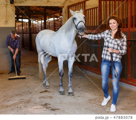 Farmer standing with horse, man cleaning floor at stabling 47883078
