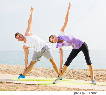 Smiling couple exercising yoga poses standing on beach Smiling couple exercising yoga poses standing on beach 47885332