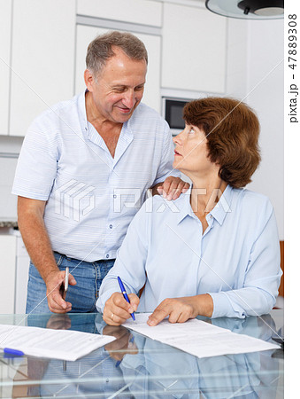Mature couple at table in home kitchen, woman filling up documents 47889308