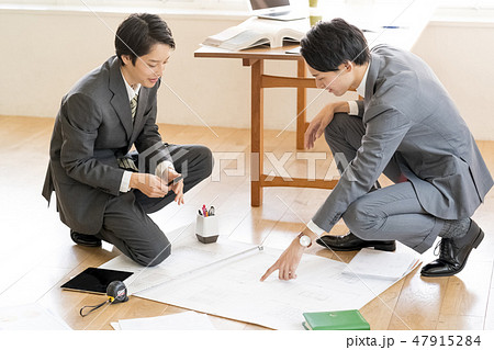 オフィスの床にしゃがみ込んで建築図面の確認をする先輩と後輩男性ビジネスマン二人 47915284